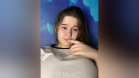 Media: Video of a young Caucasian girl with light skin and shoulder-length brown hair, wearing a white ribbed sweater. She is standing against a blue and green abstract wall, touching her nose with her index finger, smiling slightly.