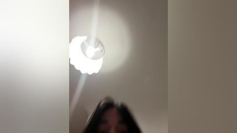 Media: A blurry video shows a woman with long, dark hair and a white ceiling light fixture with a textured shade. The image is slightly out of focus, creating a soft, dreamy effect.