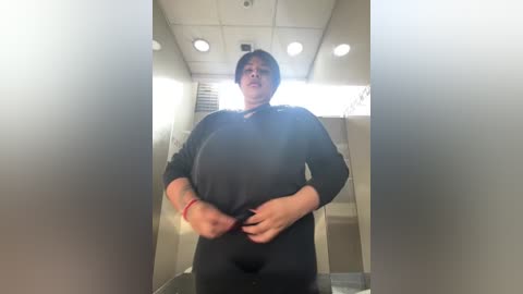 Media: Video of a short, heavyset woman with dark hair, wearing a black long-sleeve shirt and black pants, standing in a dimly lit, beige hallway with fluorescent lights and a door.