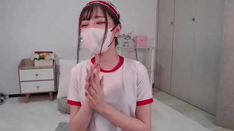 Media: Video of a young Asian woman with fair skin, wearing a white mask, red headband, and red-trimmed white t-shirt, praying in a minimalist bedroom with white walls and light-colored furniture.