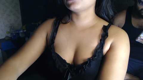 Media: A video of a light-skinned woman with long, dark hair, wearing a black, low-cut, frilled camisole that accentuates her ample cleavage, taken indoors with dim lighting.