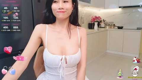 Media: Video of an East Asian woman with long black hair, wearing a white spaghetti-strap tank top, smiling in a modern kitchen.