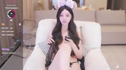 Media: Video of a slim, Asian woman with long black hair, wearing bunny ears and a black bikini, sitting on a white couch in a modern living room.