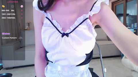 Media: Video of an Asian woman in a white, frilly maid outfit, featuring a black bow, taken indoors in a modern living room.