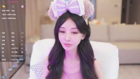 Media: Video of an East Asian woman with long black hair, wearing a white and pastel bow headband and a gray checkered top, seated indoors.