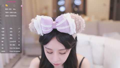 Media: A video of an Asian woman with long black hair wearing a large, frilly, pastel bow headband with furry ears, in a softly lit living room.