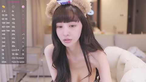 Media: A young, slender Asian woman with long black hair, wearing a teddy bear headband, sits on a couch in a modern living room. She has fair skin and is topless, revealing small breasts.