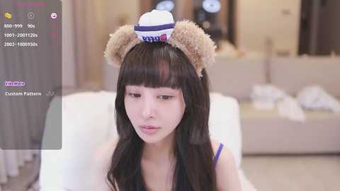 Media: Video of an East Asian woman with long black hair, wearing a white sailor hat with bear ears, and a blue top. She is indoors, sitting on a white sofa, with a blurred background.