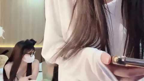 Media: Video of an Asian woman with long, straight black hair being styled by a hairstylist. She wears glasses, a white mask, and a white top. Background features a beige wall and a plush toy.