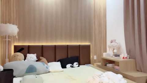 Media: Video of a modern, plush bedroom with beige walls, a plush teddy bear, and a fluffy white cat on a bed with white bedding. The room features a wooden headboard and soft lighting.