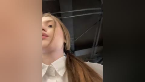 Media: Video of a young woman with fair skin and long brown hair in pigtails, wearing a white shirt, partially obscured by a person's arm on the left. Background shows industrial elements and scaffolding.