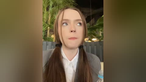 Media: A video of a young woman with fair skin, long brown hair in pigtails, wearing a white shirt and grey blazer, standing indoors with lush green plants in the background.