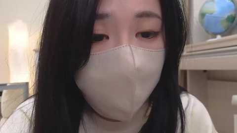 Media: Video of an Asian woman with long black hair, wearing a white surgical mask, gazing directly at the camera. She's indoors with a beige wall and a globe on a shelf in the background.