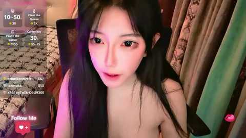 Media: Video of an East Asian woman with long black hair, pale skin, and large eyes, wearing a pink shirt, seated on a chair in a dimly lit room.