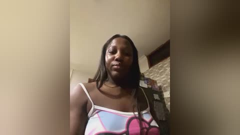 Media: Video of a dark-skinned woman with long, straight hair, wearing a colorful, patterned top, standing in a dimly lit room with beige walls and a stone wall in the background.