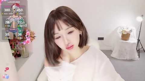 Media: A video of an East Asian woman with straight, shoulder-length brown hair and fair skin, wearing a white off-shoulder top, in a minimalist, white-walled room with a floral arrangement and a standing lamp.