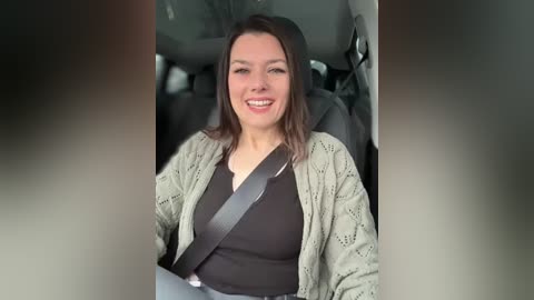 Media: Video of a smiling, light-skinned woman with shoulder-length dark hair, wearing a black top and light gray knit cardigan, sitting in a car with a seatbelt.