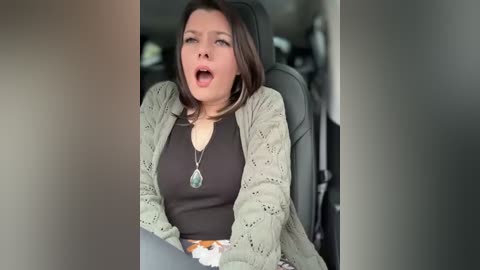 Video of a woman with short dark hair, open mouth, and surprised expression, wearing a green cardigan over a black top, seated in a car.