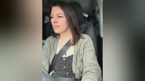 Media: Video of a woman with straight brown hair, wearing a grey cardigan over a black polka-dotted top, buckled into a car seat, looking contemplative. The background shows a blurred interior with dark upholstery.