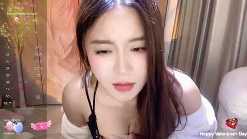 Media: Video of a young Asian woman with long brown hair, light skin, and a slender build, wearing a black bra and white shirt, in a cozy, dimly lit room with a Valentine's Day-themed background.