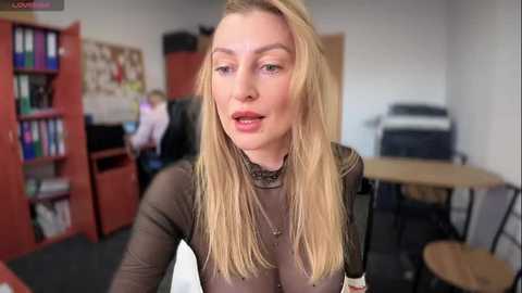 Media: Video of a blonde woman with straight hair, wearing a sheer black top, in a cluttered office with shelves, a printer, and a person in the background.