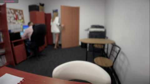 Media: Video of a modern, minimalist office with white walls and black carpet. A white chair sits in the foreground, while a woman in a white dress stands near a wooden door. Red bookshelves and a printer are visible in the background.