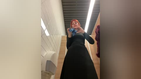 Media: Video of a pregnant woman with red hair in a black dress, taking a selfie in a public restroom with white tiled walls and a wooden floor, fluorescent lighting, and a red jacket hanging on the wall.