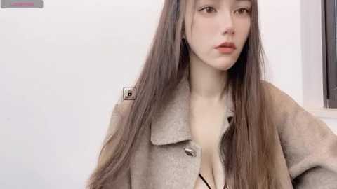 Media: Video of an Asian woman with long, straight brown hair, fair skin, and light makeup. She wears a beige, oversized cardigan and is standing against a plain white wall with a window on the right.