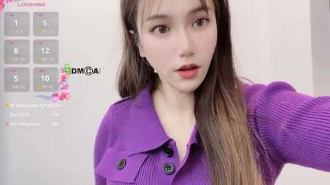 Media: Video of a young East Asian woman with fair skin and long brown hair, wearing a purple knitted jacket, in front of a white wall. Social media icons are visible in the upper left corner.