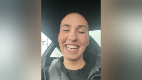 Media: Video of a smiling, light-skinned woman with a bald head, wearing a black jacket, sitting in a car with the window partially open.