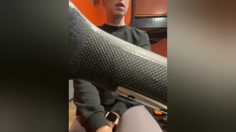 Media: Video of a person wearing a black sweatshirt and gray leggings, holding a gray, textured leg brace in their right hand, seated in a kitchen with orange walls and wooden flooring.