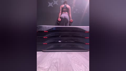 Media: Video of a woman in a sports bra and leggings performing squats on a stack of black weights, with a starry background and light wooden floor.