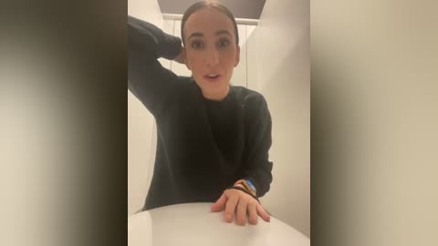 Media: Video of a young woman with light skin and dark hair, wearing a black sweater, sitting in a white, modern bathtub, looking surprised and slightly confused.