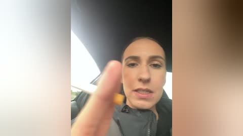 Media: Video of a light-skinned woman with short dark hair, wearing a black jacket, holding a lit cigarette. She's inside a car with a bright, sunlit window visible. The image has a grainy, low-resolution quality.