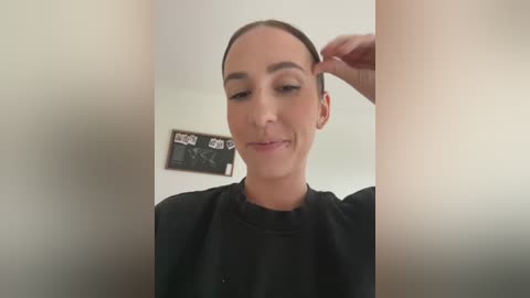 Media: Video of a fair-skinned woman with straight, dark hair, wearing a black robe, adjusting her hair in a mirror; background shows a plain white wall with a black chalkboard displaying \"Wigs\" and \"Brows.\