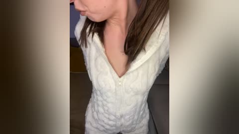 Media: A close-up video of a woman with shoulder-length brown hair, wearing a white, textured, zip-up dress, seen from the chest down, in a dimly lit room with beige walls and a wooden floor.