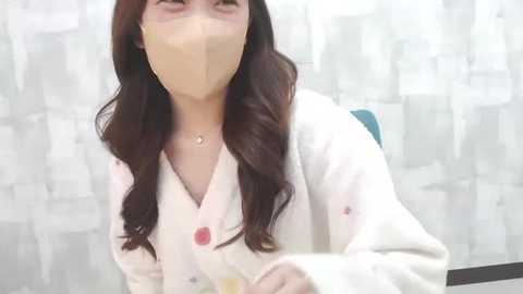 Media: Video of a smiling woman with long dark hair, wearing a beige face mask and white lab coat with pink buttons, seated in a medical or laboratory setting with white curtains in the background.