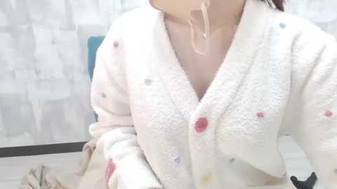 Media: Video of a woman in a white, fluffy, patterned robe with a pink button, wearing a clear face mask. She sits on a wooden chair in a light-colored room with white, textured wallpaper.