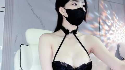 Media: Video of an East Asian woman with long black hair, wearing a black lace bra, face mask, and choker, seated on a white chair in a modern, grey-marbled room.