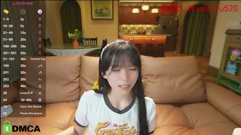 Media: A video of an East Asian woman with long black hair, wearing a white t-shirt, sitting on a beige couch in a cozy living room.