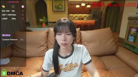 Media: Video of a young Asian woman with long black hair, wearing a white t-shirt, sitting on a beige couch in a cozy living room with wooden cabinets and a TV.