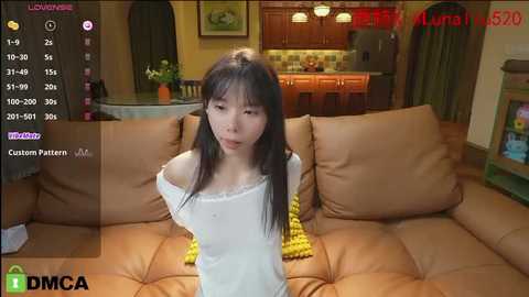 Media: A video of a young Asian woman with long black hair and fair skin wearing a white off-shoulder top, sitting on a brown leather couch in a cozy kitchen.