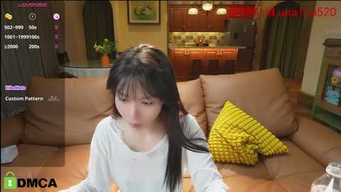 Media: A video of a young Asian woman with long black hair, wearing a white long-sleeve shirt, sitting on a brown couch in a cozy living room with wooden cabinets and a kitchen visible in the background.