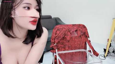 Media: Video of a young Asian woman with pale skin and shoulder-length black hair, wearing red lipstick and a purple bra, sitting on a couch. She holds a red, floral-patterned handbag with a clear plastic top. The background includes a grey couch and a yellow object.