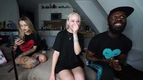 Media: A candid video of three people: a tattooed blonde in a black shirt, a blonde in a black top, and a Black man in a black hat and shirt, watching TV.