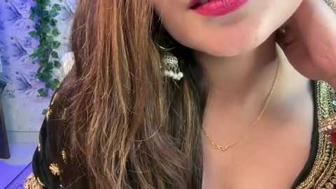 Media: Video of a woman with long, wavy brown hair, wearing a black blouse with gold embroidery, and bright pink lipstick. She has a delicate gold necklace and is indoors with a floral wallpaper background.