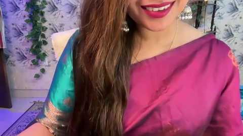 Media: Video of a smiling South Asian woman in a vibrant sari with teal and magenta colors, wearing silver earrings. Background features intricate floral wallpaper and a green vine.