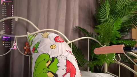 Media: Video of a green plush toy, possibly Grinch, wearing a white Santa hat, resting on a white metal bed frame with a pink hand grip. Background features green potted plants and a gray curtain.