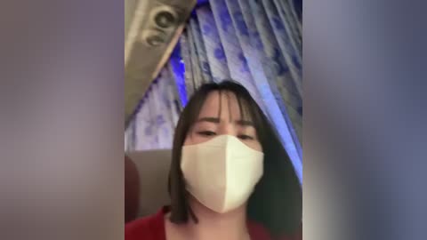 Media: A video of a woman with short black hair and a beige face mask, standing indoors, with blue and white patterned curtains in the background.