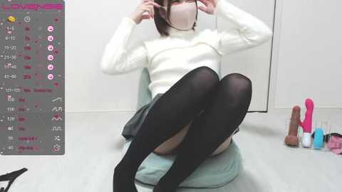 Media: Video of an Asian woman with light skin, wearing a white turtleneck, black thigh-high stockings, and a mask, seated on a light blue chair. Background includes a gray screen with text and a pink vibrator on the floor.